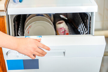 The girl closes the white dishwasher before washing the dirty dishes.