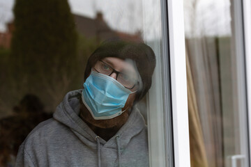 Young male looking out of window with face mask on. Lock down. Quarantine
