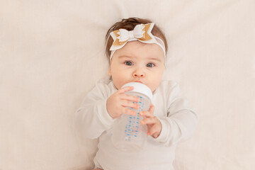 baby six months lies in a crib with a bottle and drinks water in a white bodysuit