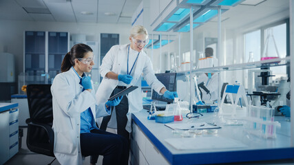 Modern Medical Research Laboratory: Two Female Scientists Working Using Digital Tablet, Analysing Biochemicals Samples. Scientific Lab for Medicine, Microbiology Development. Advanced Equipment