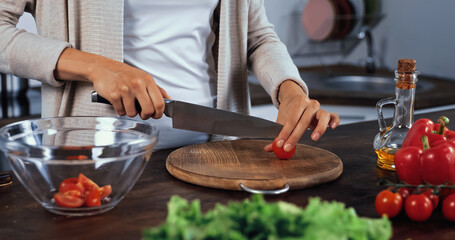 Cropped view of woman cutting cherry tomato near oil and vegetables on blurred foreground