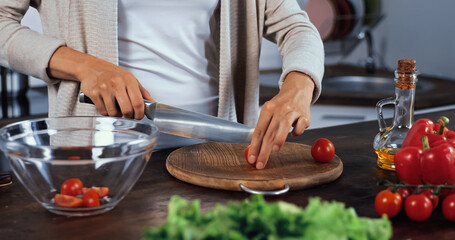 Cropped view of woman cutting cherry tomato near vegetables and lettuce on blurred foreground