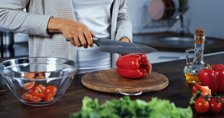 Cropped view of woman cutting fresh bell pepper near oil, lettuce and bowl on blurred foreground