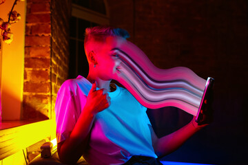 Cinematic portrait of stylish woman in neon lighted interior using a smartphone. The face is smeared, sucked, absorbed into the screen. Concept of social network dependency, phone addiction.