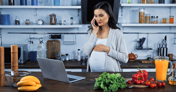 Pregnant woman talking on smartphone near laptop and food in kitchen