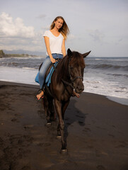 Fototapeta na wymiar Beautiful woman riding horse on the beach. Outdoor activities. Caucasian woman wearing jeans and white T-shirt. Traveling concept. Cloudy sky. Bali, Indonesia