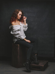 A girl with red and curly hair in a white shirt on a dark background. A girl in jeans sits on a wooden crate.