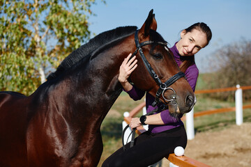 Young female farmer taking care about horse on ranch on summer day, copy space. Girl stroking and hugging brown horse
