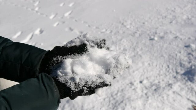 Hands in black gloves hold white snow and try to sculpt a snowball on the background of a winter sunny day.