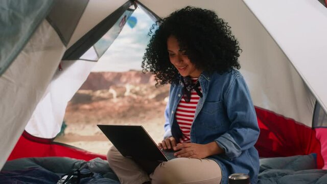 Excited Beautiful Young Multiethnic Female Traveler with Afro Hairstyle Using Laptop Computer in a Tent on Top of Rocky Canyon