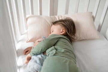 child sleeps on its side in an olive-colored suit, with a toy rabbit for sleeping, in a white crib