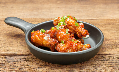 Chicken stir-fried with spicy sauce sprinkled with white sesame and chopped scallions is placed in a black ceramic pan on a wooden table.