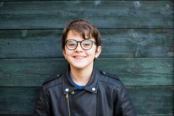 portrait of 12 year old boy with glasses smiling on green wood background dressed in leather jacket