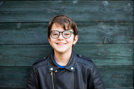 portrait of 12 year old boy with glasses smiling on green wood background dressed in leather jacket