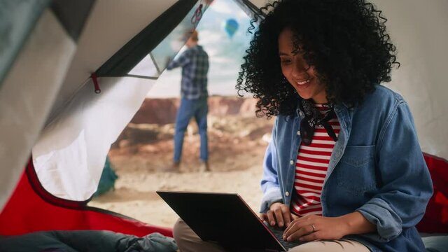 Diverse Adventurous Couple Mountain Hikers Living in Nature in Great Outdoors. Multiethnic Female Traveller with Afro Hairstyle Using Laptop Computer in a Tent on Top of Rocky Canyon. 
