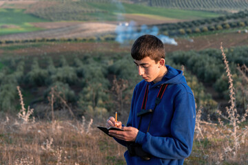 Young boy riting in little notebook while admiring beautiful landscape in the countryside.