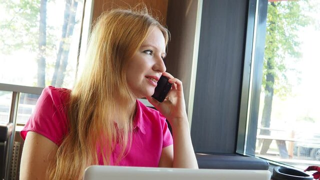 A young attractive business woman blonde in a pink shirt sitting in a cafe with a laptop and coffee. works at a computer, communicates on the phone, writes messages. freelance