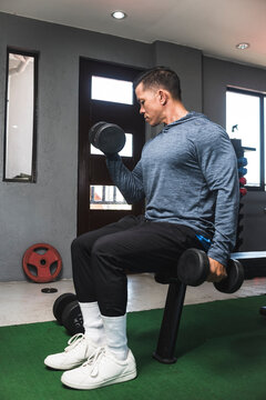 A Fit Athletic Asian Guy In A Sweatshirt Does Seated Alternate Dumbbell Curls. Working Out And Training Biceps. Open Air Gym Setting.