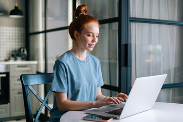 Side view of smiling redhead young woman working typing on laptop computer sitting at table in kitchen room. Concept of remote working from home office during quarantine from COVID-19.
