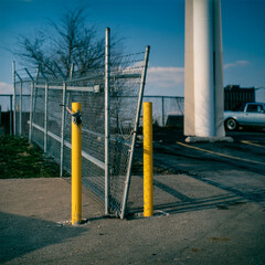 A minimal abstract urban detail study of two yellow parking bollards protecting a gate of a fence in a parking lot with a large signpost in the background.  Kansas City Missouri 1985