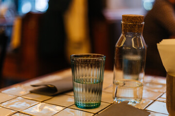 Corrugated clear tall glass of water stands on served table (cutlery and napkin) next to trendy jug in a restaurant. Blurred people on background