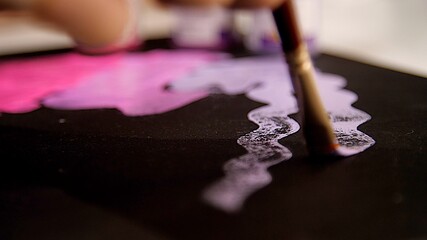 Artist's hand holds a paintbrush and draws a wavy line of pink or lilac on the black canvas. Close view. Pink color of oil paint or watercolor. Acrylic drawing concept, artist work