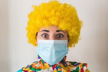 Linda mujer joven sonriendo mientras está vestida con un disfraz de payaso con una mascarilla quirúrgica desechable y una peluca de payaso amarilla. Protección contra el virus sars-cov-2 - obrazy, fototapety, plakaty