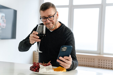 Happy adult unshaven man using mobile phone while drinking wine