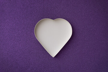 An empty white heart shaped box on a purple glittering background. 