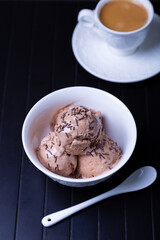Chocolate ice cream with chocolate sprinkle in a white bowl on a black background. Three balls of homemade ice cream and a cup of coffee. Close-up.