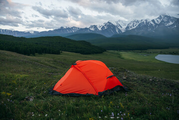 Atmospheric alpine landscape with orange tent on background of lake and big snowy mountains in overcast weather. Awesome green scenery with orange tent with view of great mountains under cloudy sky.