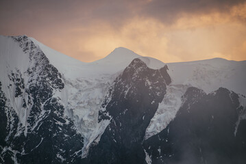 Minimal alpine landscape with snowy mountain top among low clouds on background of sunset or sunrise cloudy sky of illuminating color. Atmospheric scenery with snow-white mountain ridge in dawn sky.