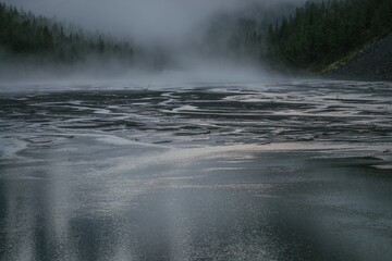 Atmospheric alpine scenery with mountain lake and water streams on background of forest silhouette in low clouds. Scenic highland landscape with mountain streams and trees silhouettes in fog on dusk.