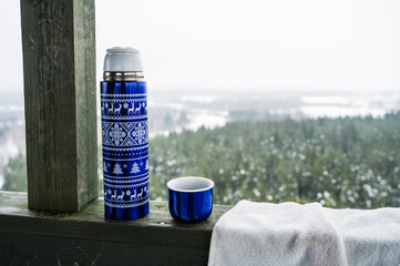 Close-up of blue thermos bottle with patterns on wooden surface. Blurred winter forest on background. Outdoor picnic.