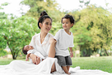 Asian young beautiful mother holding her newborn is sleeping and feel with love and touching gently then sitting on green grass in the park