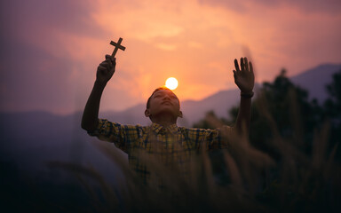 Boy holding a Cross overhead for pray to God with light sunset background, beliefs of children...