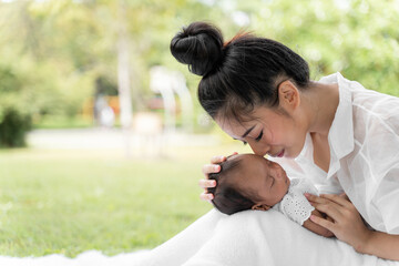 Asian young beautiful mother holding her newborn is sleeping and feel with love and touching gently then sitting on green grass in the park