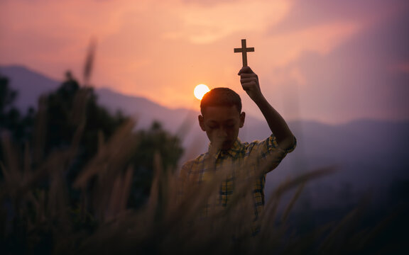 Christian children holding a Cross overhead for pray to God with light sunset background, beliefs of children concept.