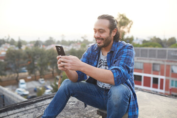 30-year-old man sitting on his back on the rooftop of a building watching the sunset and browsing on his smart cell phone