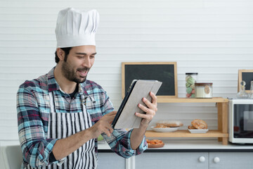 Happy handsome Caucasian man with beard wear chef hat and apron smile holding using tablet in the kitchen