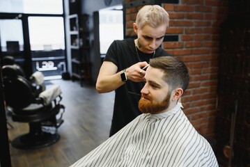 Making haircut look perfect. Young bearded man getting haircut by hairdresser while sitting in chair at barbershop