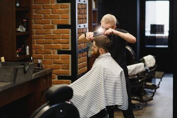 Making haircut look perfect. Young bearded man getting haircut by hairdresser while sitting in chair at barbershop