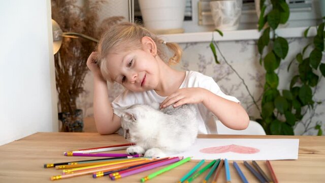 a little girl is stroking a white kitten that is playing with colored pencils on a wooden table
