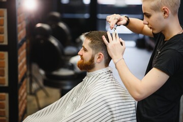 Making haircut look perfect. Young bearded man getting haircut by hairdresser while sitting in...