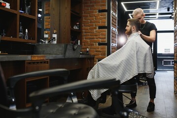 Making haircut look perfect. Young bearded man getting haircut by hairdresser while sitting in...