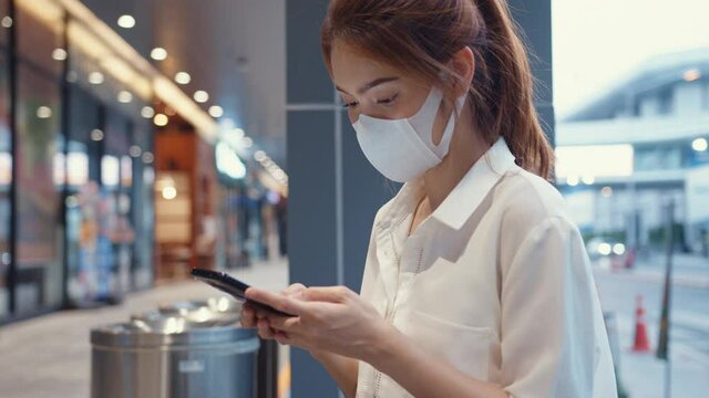 Young Asia businesswoman in fashion office clothes wearing medical face mask using smart phone typing text message while sitting outdoors in urban modern city at night. Business on the go concept.