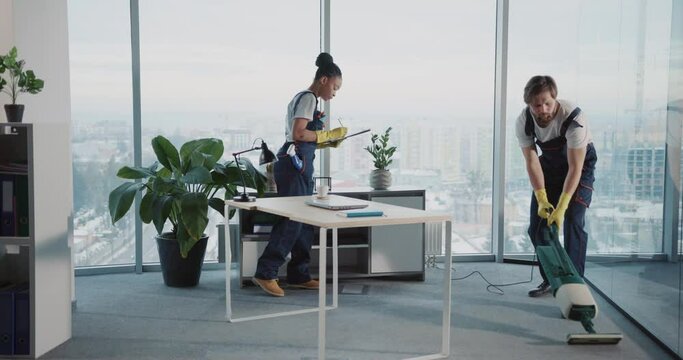 Office cleaning. Young male worker using vacuum cleaner tidying corporate workspace. Female colleague african woman walking around office room checking writing notes.