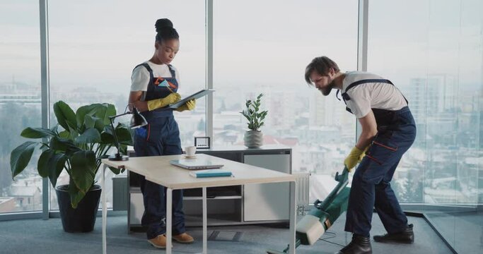 Multi-ethnic couple of janitors cleaning corporate workspace office room with vacuum cleaner cooperating together. Cleaning services. Modern interiors.