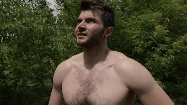 Sweaty bearded guy with a bare hairy chest rests after sports looking at the sun and exhaling air