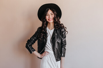 Asian girl in leather jacket standing with hand on hip. Studio shot of smiling chinese woman in hat isolated on beige background.